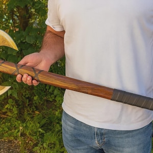 Könnte beinhalten: Eine zweischneidige Axt mit Holzgriff und goldfarbenen Klingen. Der Griff ist am Ende mit dunkelgrauem Material umwickelt. Die Axt wird vor einer Person gehalten, die ein weißes T-Shirt und eine blaue Jeans trägt.