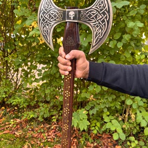 May include: A double-bladed axe with a dark wooden handle and a silver-colored head. The head features intricate Celtic knotwork and a skull emblem. The handle is adorned with gold-colored floral patterns. The axe is held against a backdrop of green foliage.