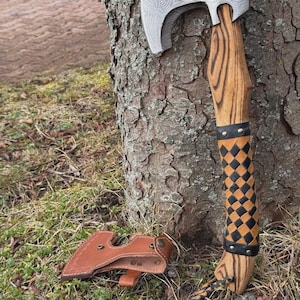 May include: Axe with a silver blade featuring an engraved design, a wooden handle, and a black and orange checkered grip. A brown leather sheath rests on the ground near the axe.