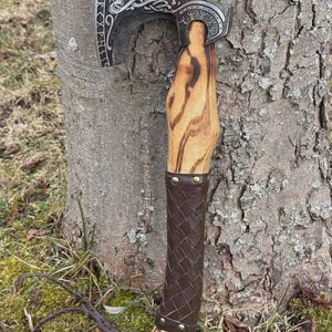 May include: Axe with a silver blade featuring engraved designs and text. The wooden handle has a brown leather-wrapped grip. The axe is leaning against a tree trunk, showcasing its craftsmanship and rustic aesthetic.