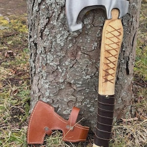May include: A handcrafted axe with a polished steel blade and a wooden handle. The handle is wrapped in dark brown leather and features a decorative carved design. A brown leather sheath protects the blade. The axe is leaning against a tree trunk.