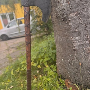 Puede incluir: Un hacha artesanal con una hoja oscura y texturizada y un mango de madera. El mango est&aacute; envuelto con un material similar al cuero y presenta un patr&oacute;n de cruz decorativo cerca de la cabeza. El hacha est&aacute; apoyada contra un tronco de &aacute;rbol.