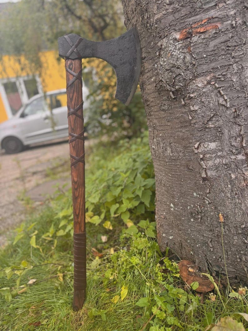 Puede incluir: Un hacha artesanal con una hoja oscura y texturizada y un mango de madera. El mango est&aacute; envuelto con un material similar al cuero y presenta un patr&oacute;n de cruz decorativo cerca de la cabeza. El hacha est&aacute; apoyada contra un tronco de &aacute;rbol.