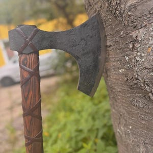 Puede incluir: Un hacha artesanal con una hoja oscura y texturizada y un mango de madera. El mango est&aacute; envuelto con tiras de cuero marr&oacute;n. El hacha est&aacute; apoyada contra un tronco de &aacute;rbol, mostrando su dise&ntilde;o r&uacute;stico.