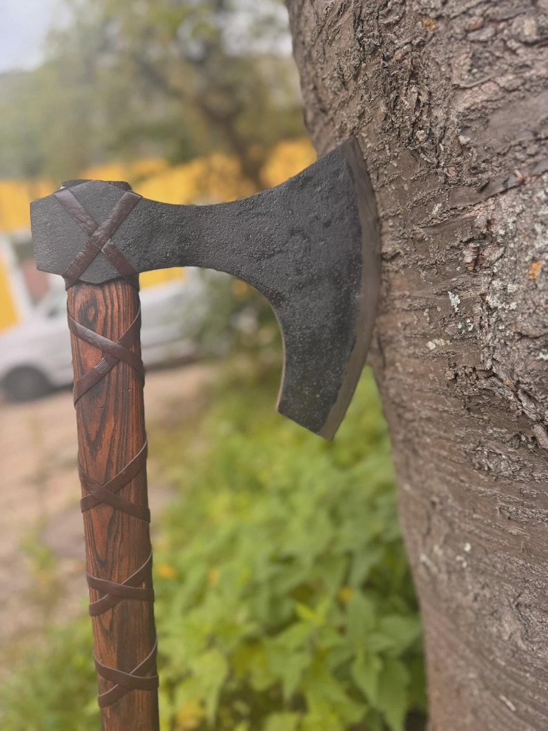 Puede incluir: Un hacha artesanal con una hoja oscura y texturizada y un mango de madera. El mango est&aacute; envuelto con tiras de cuero marr&oacute;n. El hacha est&aacute; apoyada contra un tronco de &aacute;rbol, mostrando su dise&ntilde;o r&uacute;stico.