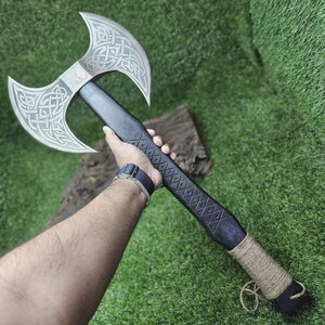 May include: A double-bladed axe with a black handle and silver blades. The blades feature intricate Celtic knot designs and a skull emblem. The handle is wrapped with twine near the base, and the axe is displayed against a green background.