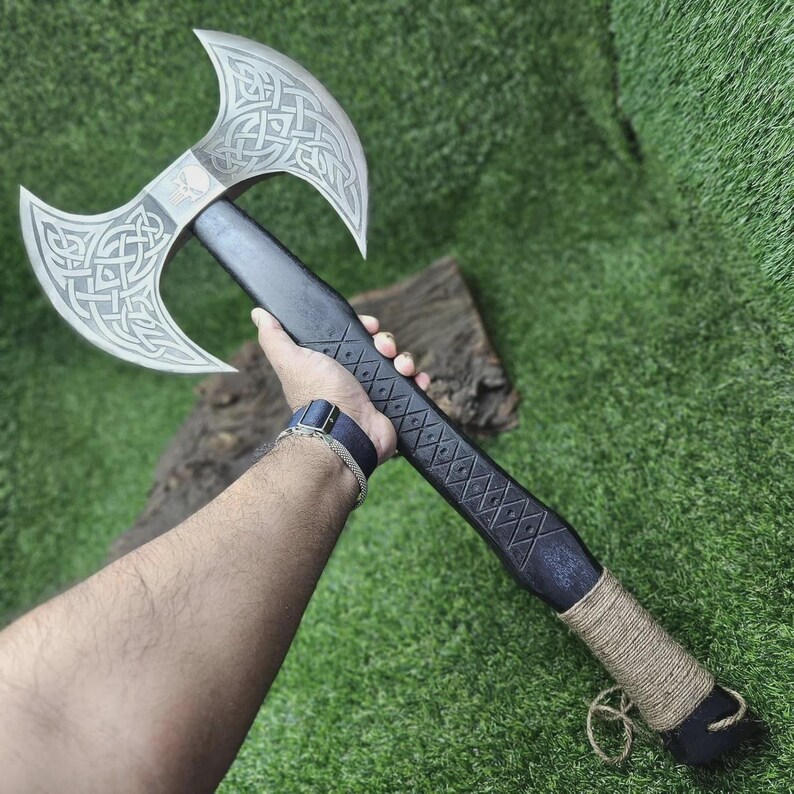 May include: A double-bladed axe with a black handle and silver blades. The blades feature intricate Celtic knot designs and a skull emblem. The handle is wrapped with twine near the base, and the axe is displayed against a green background.