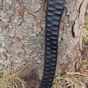 May include: A close-up of a black, carved wooden handle with a textured, wave-like pattern. The handle is positioned vertically against a tree trunk with rough, brown bark. The handle's design suggests a tool or weapon.