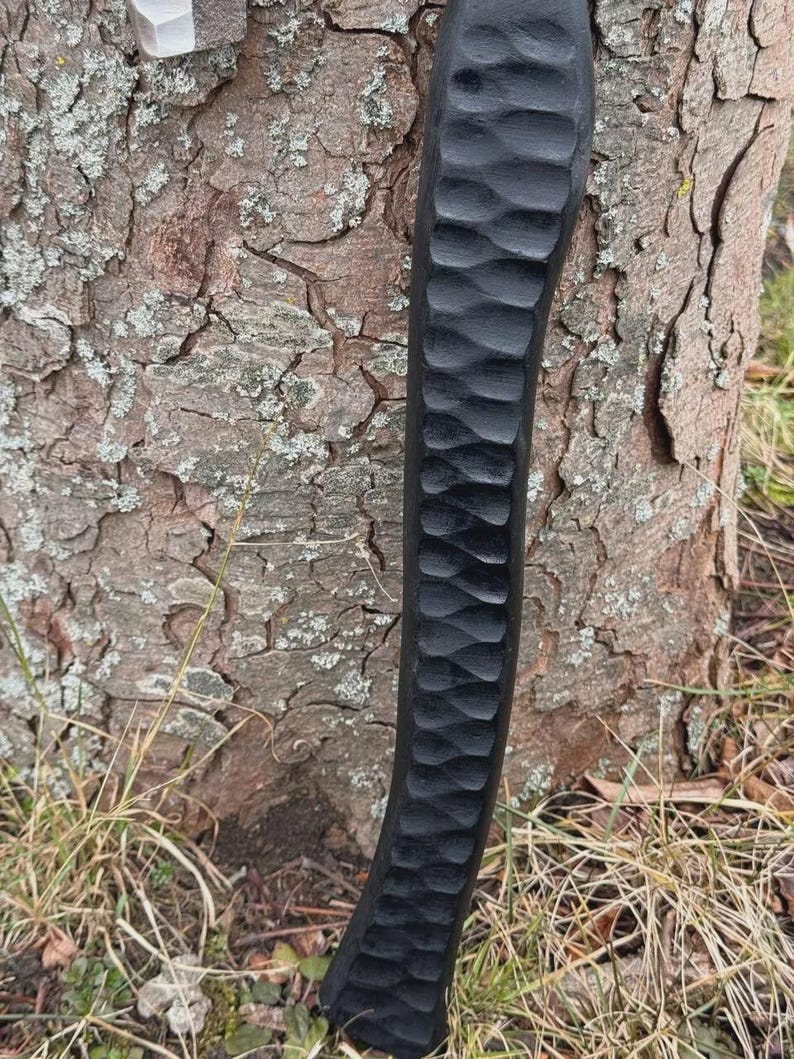 May include: A close-up of a black, carved wooden handle with a textured, wave-like pattern. The handle is positioned vertically against a tree trunk with rough, brown bark. The handle's design suggests a tool or weapon.