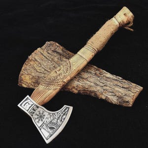 May include: A handcrafted axe with a wooden handle and a silver-colored blade. The handle is intricately carved with geometric patterns and wrapped with twine. The blade features etched designs, including a symbol resembling a compass. The axe rests on a piece of wood.