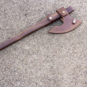 May include: A traditional axe featuring a dark wooden handle and a brown leather sheath. The axe head is metal with a curved blade. The leather sheath is secured with metal rivets and measures approximately 25 cm.