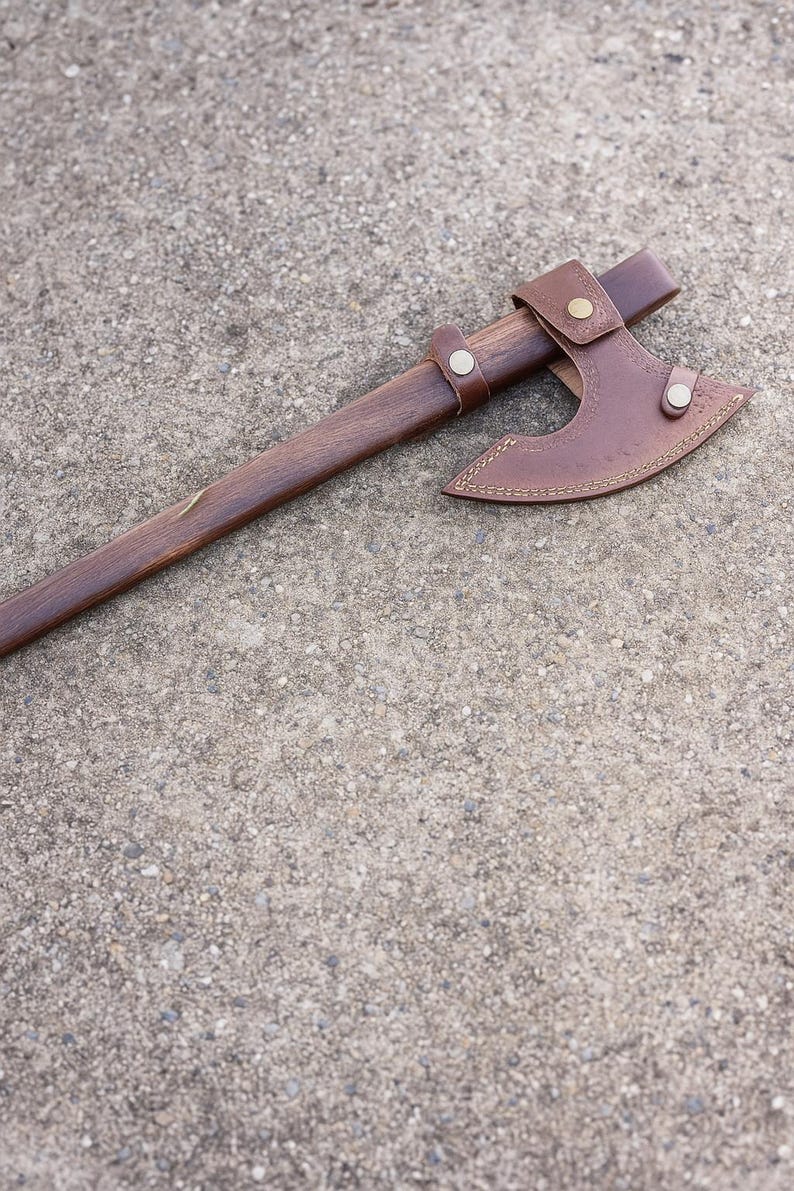 May include: A traditional axe featuring a dark wooden handle and a brown leather sheath. The axe head is metal with a curved blade. The leather sheath is secured with metal rivets and measures approximately 25 cm.