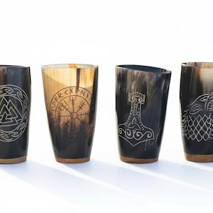 Può includere: Quattro bicchieri di corno vichinghi con disegni incisi. Le tazze presentano simboli come il Valknut, Vegvisir, Mjolnir e un lupo. Ogni tazza ha una base marrone scuro e un colore di corno naturale.