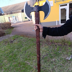 May include: A large, double-bladed axe with a dark metal head and a long, wooden handle. The handle is wrapped with brown leather and features decorative cross-stitch patterns. The axe is held up against a background of green grass and a building.