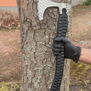 May include: A silver and black axe with a textured handle and blade. The axe head is silver, and the handle is black. The axe is held against a tree trunk. The axe is approximately 15 inches long.