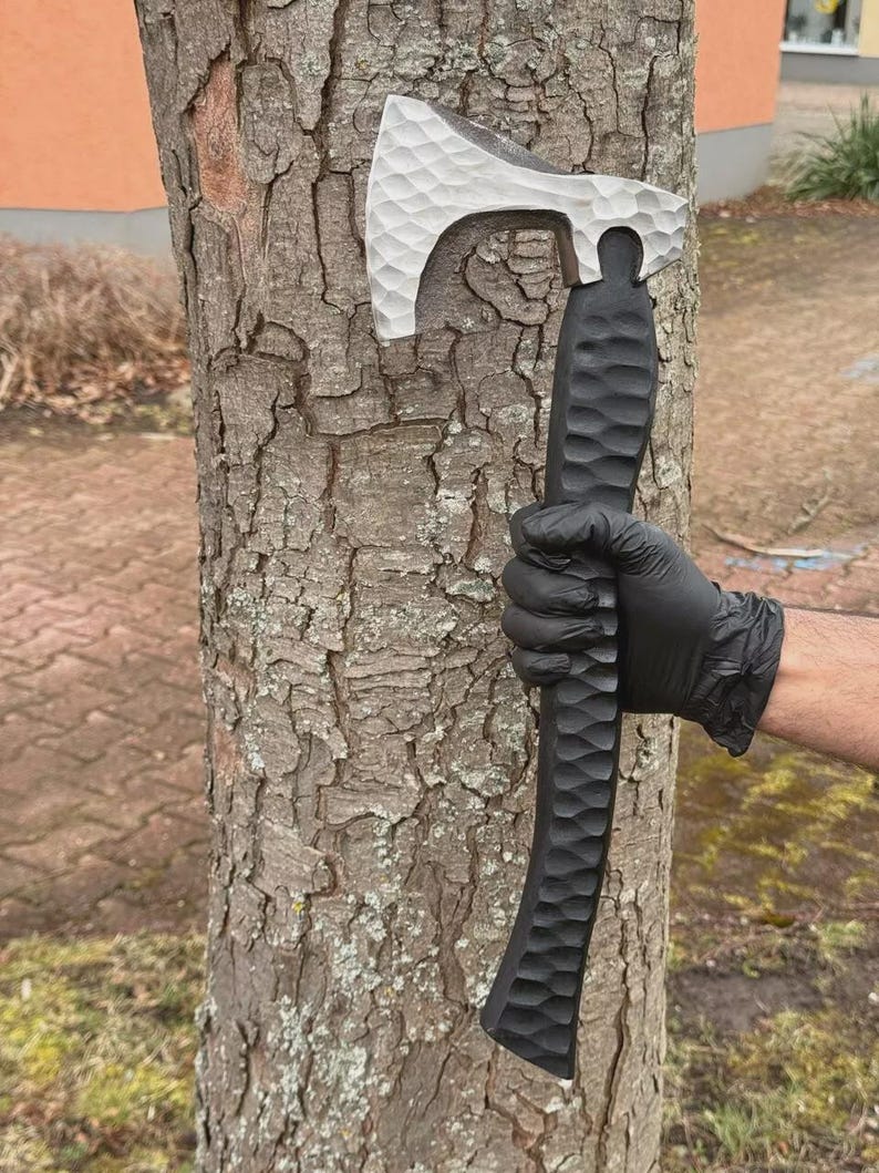 May include: A silver and black axe with a textured handle and blade. The axe head is silver, and the handle is black. The axe is held against a tree trunk. The axe is approximately 15 inches long.