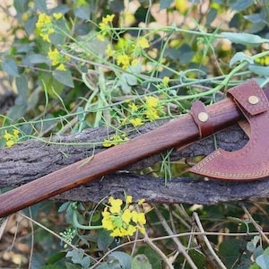 May include: A handcrafted axe with a wooden handle and a leather sheath. The axe head is made of metal and has a brown leather cover. The axe is resting on a piece of wood, surrounded by greenery.