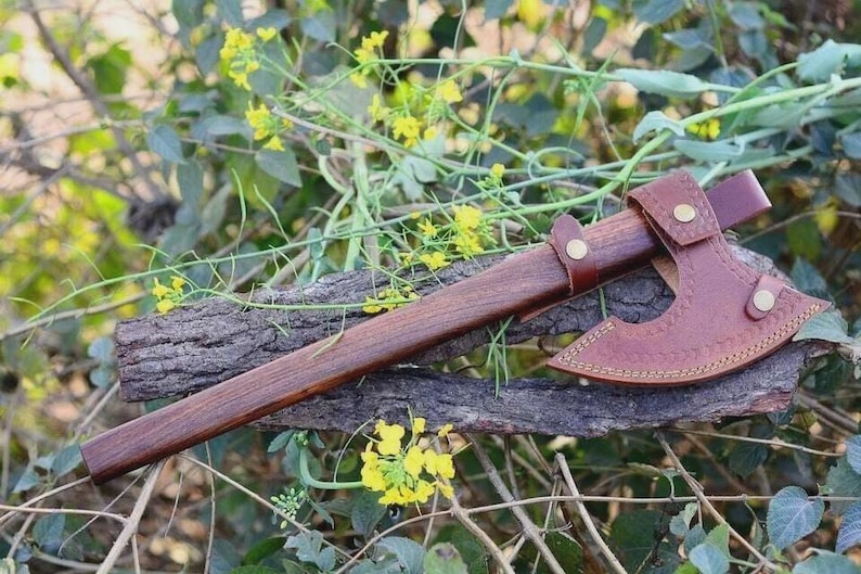 May include: A handcrafted axe with a wooden handle and a leather sheath. The axe head is made of metal and has a brown leather cover. The axe is resting on a piece of wood, surrounded by greenery.
