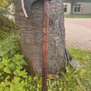 Könnte beinhalten: Eine Axt im Wikinger-Stil mit einer dunklen, gebogenen Klinge und einem Holzgriff. Der Griff ist mit Lederbändern im Kreuzmuster umwickelt. Die Axt lehnt an einem Baumstamm und zeigt ihr rustikales Design und ihre Handwerkskunst.
