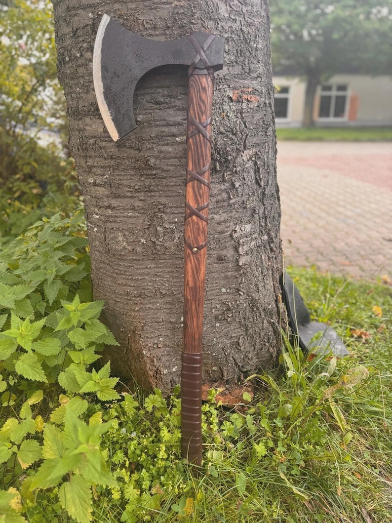 Puede incluir: Un hacha de estilo vikingo con una hoja oscura y curvada y un mango de madera. El mango est&aacute; envuelto con tiras de cuero en un patr&oacute;n entrecruzado. El hacha est&aacute; apoyada contra un tronco de &aacute;rbol, mostrando su dise&ntilde;o r&uacute;stico y artesan&iacute;a.