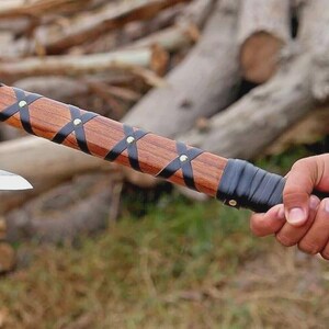 Puede incluir: Un hacha artesanal con mango de madera y hoja plateada. El mango est&aacute; envuelto con correas de cuero negro y asegurado con remaches. El hacha est&aacute; sujeta en la mano de una persona.