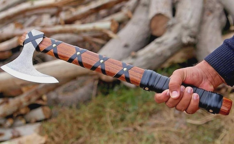 Puede incluir: Un hacha artesanal con mango de madera y hoja plateada. El mango est&aacute; envuelto con correas de cuero negro y asegurado con remaches. El hacha est&aacute; sujeta en la mano de una persona.