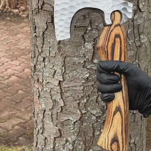 May include: A handcrafted axe with a textured, silver-colored head and a wooden handle. The handle features a unique wood grain pattern. The axe is held against a tree trunk.
