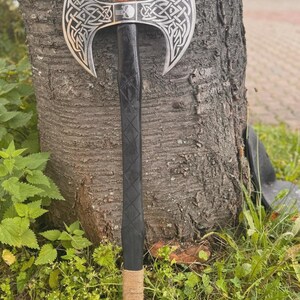 Puede incluir: Un hacha de doble hoja con una cabeza plateada que presenta intrincados diseños de nudos celtas. El mango de madera negra tiene un patrón de diamantes y está envuelto con cuerda natural cerca de la base. El hacha está apoyada contra un tronco de árbol.