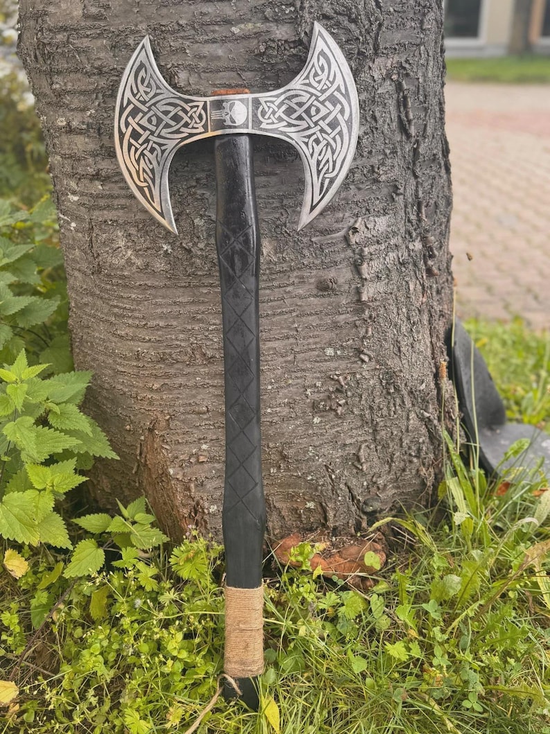May include: A double-bladed axe with a silver head featuring intricate Celtic knot designs. The black wooden handle has a diamond pattern and is wrapped with natural twine near the base. The axe is leaning against a tree trunk.