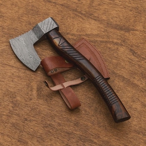 May include: A handcrafted axe with a patterned steel blade and a dark wooden handle. The handle features carved details and is paired with a brown leather sheath. The axe is designed for outdoor use and is shown on a wooden surface.