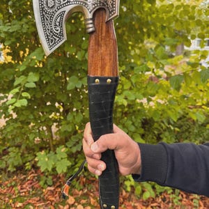 Könnte beinhalten: Axt mit einem silberfarbenen Kopf mit aufwendigen Gravuren und einem Holzgriff. Der Griff ist mit schwarzem Leder umwickelt, das mit messingfarbenen Nieten befestigt ist. Die Axt wird vor einem Hintergrund aus grünem Laub gehalten.