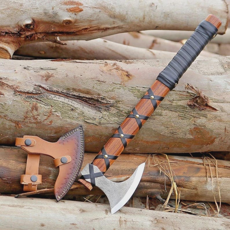 Puede incluir: Un hacha artesanal con un mango de madera envuelto en cuero negro. La cabeza del hacha es plateada con una hoja curva. Se incluye una funda de cuero marr&oacute;n. El hacha mide aproximadamente 46 cm de largo.