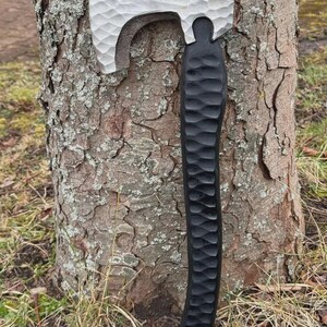May include: A handcrafted axe with a textured, silver-colored head and a black, carved wooden handle. The axe is leaning against a tree trunk, showcasing its detailed craftsmanship and rustic aesthetic.