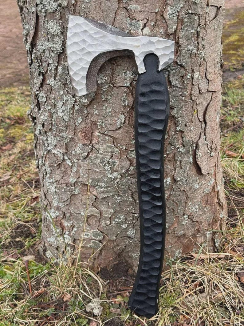 May include: A handcrafted axe with a textured, silver-colored head and a black, carved wooden handle. The axe is leaning against a tree trunk, showcasing its detailed craftsmanship and rustic aesthetic.