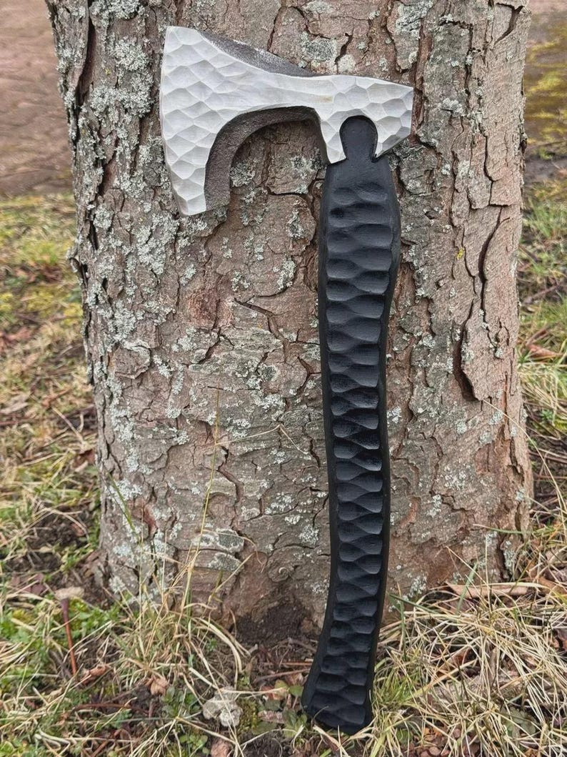 May include: A handcrafted axe with a textured, silver-colored head and a black, carved handle. The axe is leaning against a tree trunk, showcasing its detailed craftsmanship. The axe is a tool for outdoor activities.