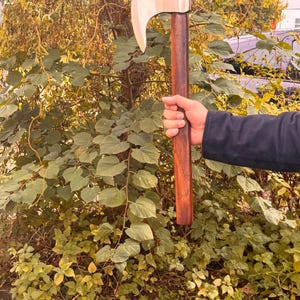 May include: A wooden axe with a light-colored blade and a dark brown handle. The axe is held in a person's hand against a backdrop of green foliage and trees. The axe appears to be new and unused.
