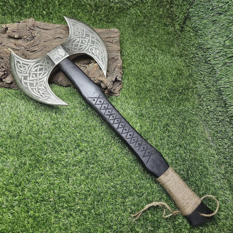 May include: A double-bladed axe with a silver-colored head featuring intricate Celtic knotwork and a skull emblem. The black handle is adorned with diamond-shaped carvings and wrapped with twine near the base. The axe rests on a piece of wood against a green background.