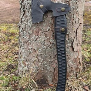 May include: A black axe with a leather sheath, resting against a tree trunk. The axe head is dark, and the handle is carved with a textured pattern. The leather sheath is secured with brass-colored rivets.