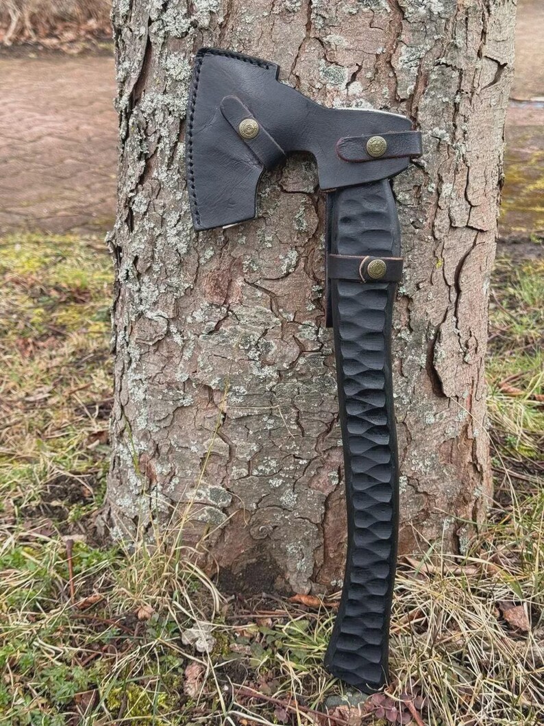 May include: A black axe with a leather sheath, resting against a tree trunk. The axe head is dark, and the handle is carved with a textured pattern. The leather sheath is secured with brass-colored rivets.