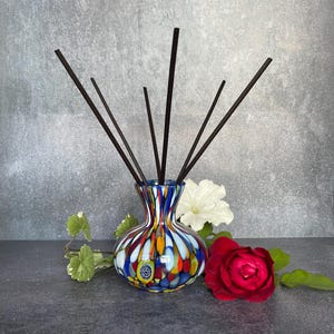 May include: A decorative glass diffuser vase with a colourful pattern of blue, red, yellow, and white. The vase contains several dark brown diffuser sticks. A red rose and white flower are placed nearby.