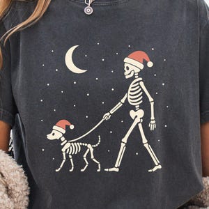 May include: Dark gray t-shirt featuring a skeleton walking a skeleton dog, both wearing Santa hats, under a crescent moon and stars. The graphic is in a cream color. A cozy, cream-colored sweater is draped over the person's shoulders.
