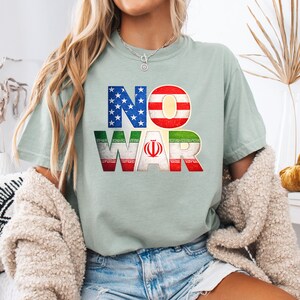 May include: A light green t-shirt with the words "NO WAR" in a bold, colorful design. The letters are filled with the American and Iranian flags. The shirt is a casual, everyday wear item.