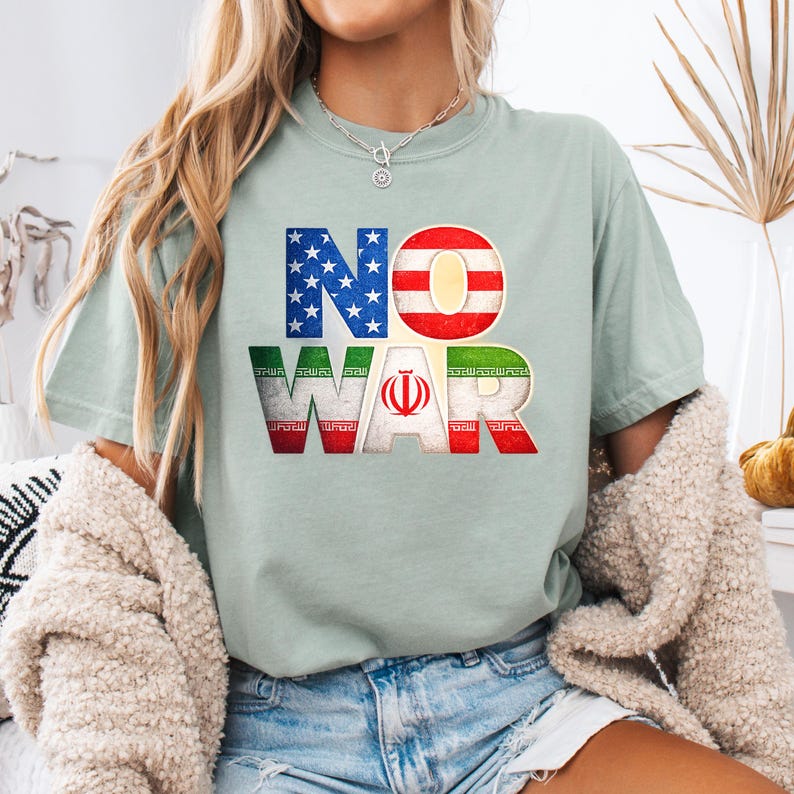 May include: A light green t-shirt with the words "NO WAR" in a bold, colorful design. The letters are filled with the American and Iranian flags. The shirt is a casual, everyday wear item.