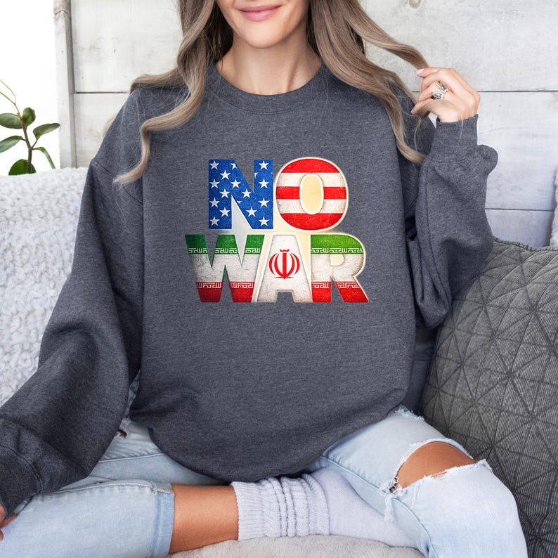 May include: Dark gray sweatshirt with the words "NO WAR" in a distressed font. The letters are designed with the American and Iranian flags. The sweatshirt is a crew neck style.