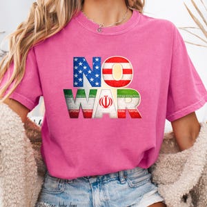 May include: A pink t-shirt with the words "NO WAR" in a bold, colorful design. The letters are filled with the American and Iranian flags. The shirt has short sleeves and a crew neck.