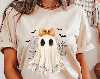 May include: Beige t-shirt featuring a cartoon ghost with a bow, spiderweb, and bats. The ghost has a white lace trim and black oval eyes. The design is perfect for Halloween or spooky season.