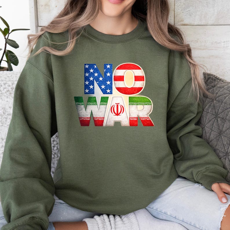 May include: An olive green sweatshirt with the words "NO WAR" in a distressed, patriotic design. The letters "NO" are in the colors of the American flag, and "WAR" is in the colors of the Iranian flag.