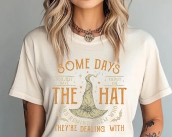May include: Cream-colored t-shirt with the text "SOME DAYS YOU JUST HAVE TO PUT ON THE HAT AND REMIND THEM WHO THEY'RE DEALING WITH." The graphic features a witch's hat in the center, with stars and bats.