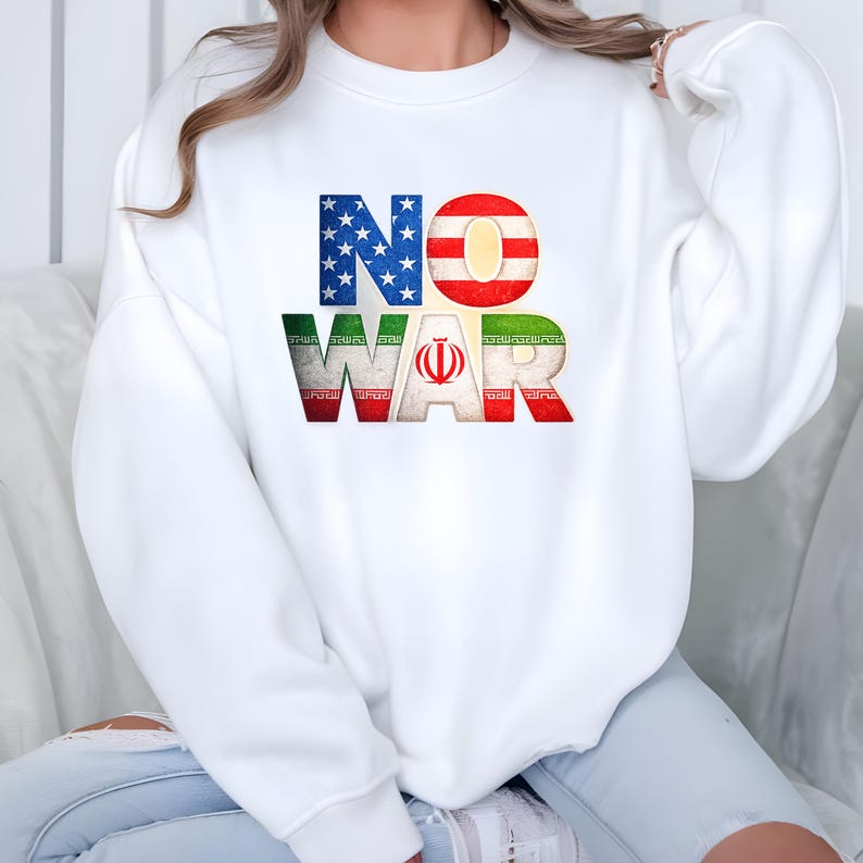 May include: White sweatshirt with the words "NO WAR" in a distressed, patriotic design. The letters are colored with the American and Iranian flags. The "N" is blue with white stars, the "O" is red and white stripes, and "WAR" is green, red, and white.