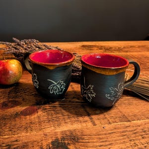 Puede incluir: Dos tazas de cerámica negra con interiores rojos y diseños de hojas. Los bordes tienen un esmalte marrón. Las tazas están sobre una superficie de madera con una manzana y plantas secas en el fondo. Cada taza tiene un asa.